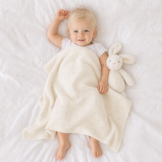 Petite fille blonde de 12 mois allongée avec une couverture bébé tricot crème à maille ajourée et un lapin en peluche.