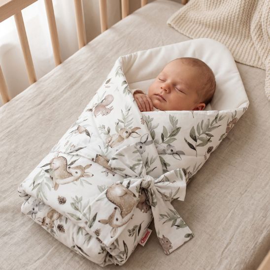 Bébé emmailloté dans une couverture motif forêt enchantée, allongé dans un lit à barreaux