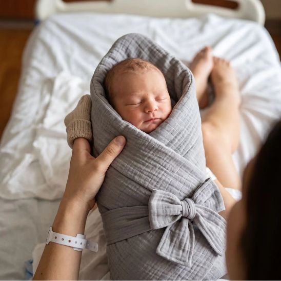 Nouveau-né emmailloté dans une couverture grise, tenu par les mains d'un parent à la maternité
