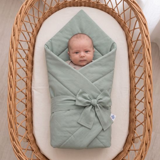Bébé emmailloté dans une couverture unie vert sauge, installé dans un couffin en osier