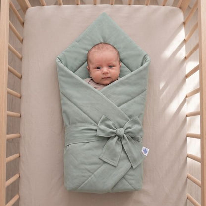 Bébé emmailloté dans une couverture unie vert sauge, allongé dans un lit à barreaux ensoleillé