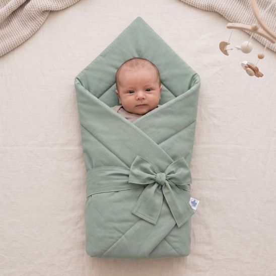 Bébé emmailloté dans une couverture unie vert sauge, posé sur du linge avec mobile en arrière-plan