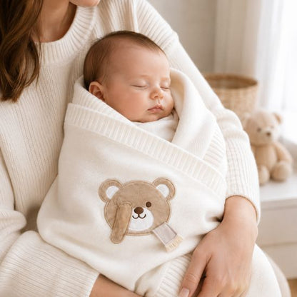 Bébé emmailloté dans une couverture tricot blanc écru avec ourson brodé, dans les bras de sa maman
