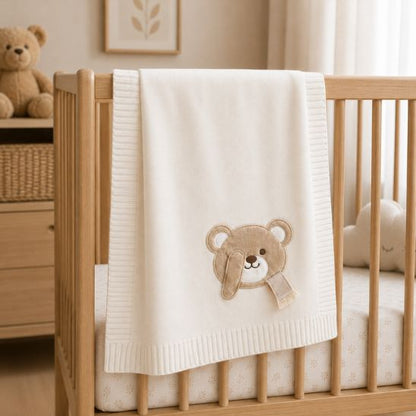 Couverture tricot pour bébé blanc écru avec ourson brodé, posée sur un lit à barreaux
