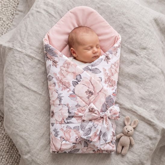 Bébé emmailloté dans une gigoteuse réversible motif fleurs roses, allongé sur un lit avec un doudou lapin en tricot