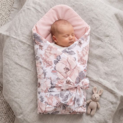 Bébé emmailloté dans une gigoteuse réversible motif fleurs roses, allongé sur un lit avec un doudou lapin en tricot