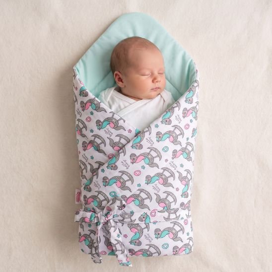 Bébé emmailloté dans une gigoteuse réversible motif chevaux à bascule, sur un drap clair