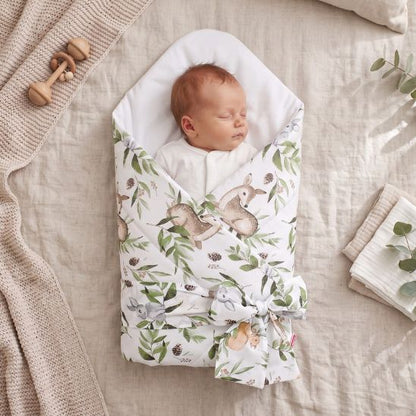 Bébé emmailloté dans une gigoteuse réversible motif forêt, posé sur du linge de maison