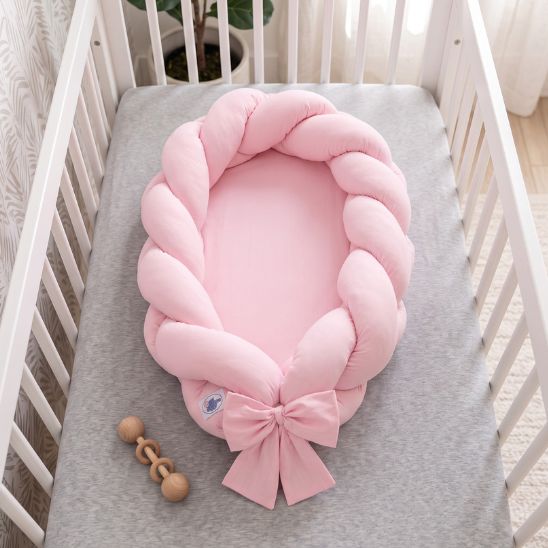 Réducteur de lit tressé rose vu de dessus dans un lit à barreaux, avec hochet en bois