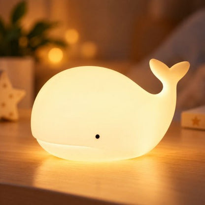 Veilleuse baleine blanche allumée en lumière chaude sur une table de chevet en bois